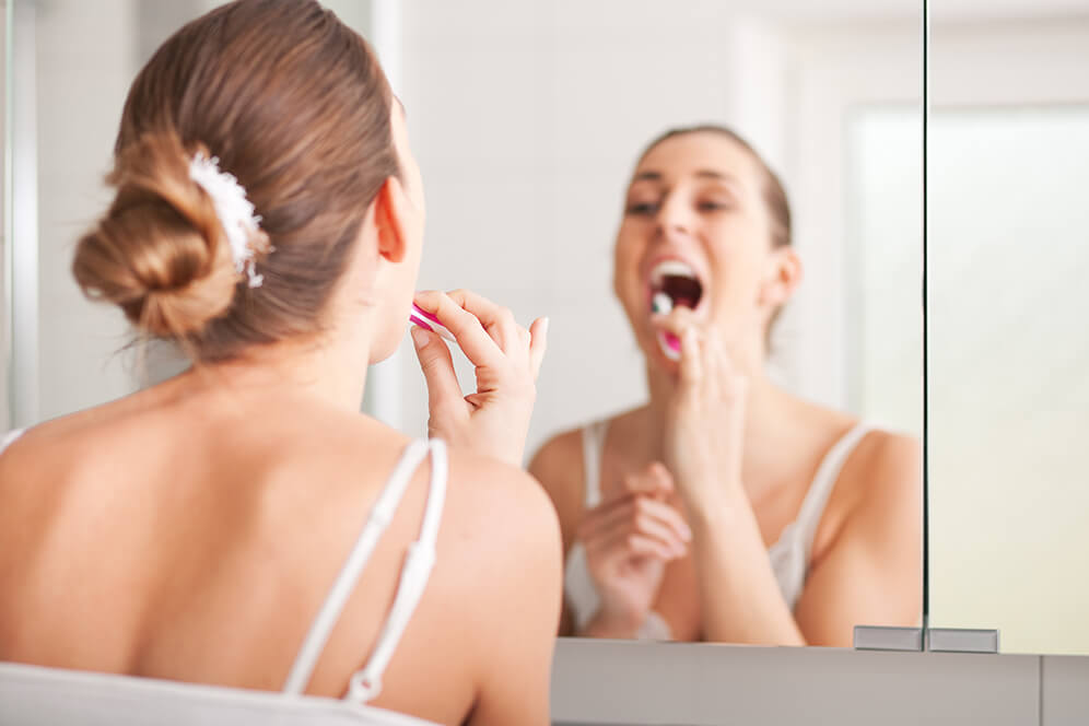 Ganzheitlichen Mundgesundheit, Hamburg - Eine Junge Dame putzt sich die Zähn und schaut im Badezimmerspiegel.